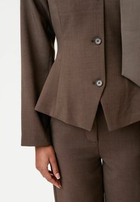 Hnedý strihový oblek s priliehavým blejzrom v peplum štýle, ktorý má dva gombíky a zladené nohavice, vyrobený z hladkej, texturovanej tkaniny.
