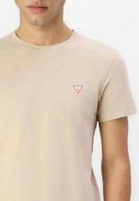 Camiseta de algodón beige con mangas cortas. Presenta un pequeño logo triangular rojo y blanco en el lado izquierdo del pecho. Textura suave y corte clásico.