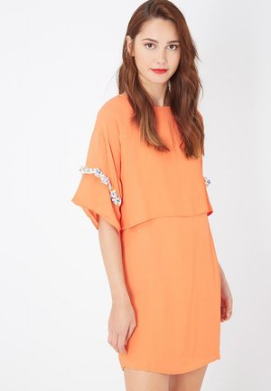 Mujer joven con cabello largo castaño rojizo, vistiendo un vestido naranja suelto de manga corta con volantes estampados en las mangas, de pie contra un fondo blanco.