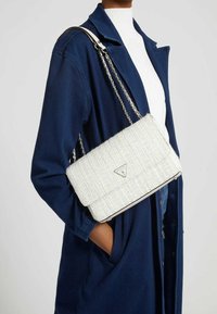 Sac à main en tweed blanc avec une forme structurée, une anse en chaîne argentée et un accent logo. Le modèle porte un manteau bleu foncé et un top blanc.