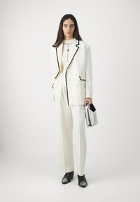 White double-breasted suit with black trim, tailored fit, paired with black ankle boots featuring silver toe caps and a small metallic clutch.