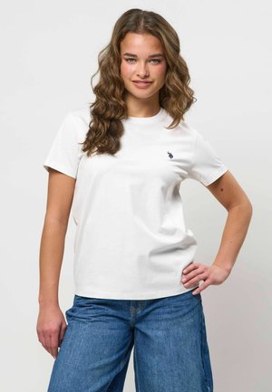 Mladá žena s vlnitými hnedom vlasmi, oblečená v bielom tričku s krátkym rukávom a modrých džínsoch, stojaca s jednou rukou na bedrách.