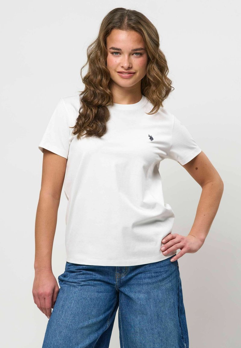 Jeune femme aux cheveux bruns ondulés portant un t-shirt blanc à manches courtes et un jean bleu, debout avec une main sur la hanche.