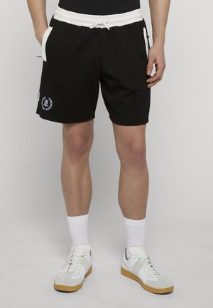 Persoon die zwarte sportshorts met een witte koordsluiting, witte sokken en witte sneakers met bruine zolen draagt, staand tegen een effen achtergrond.