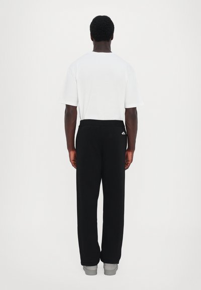 Weißes T-Shirt mit kurzen Ärmeln und schwarze Jogginghose. Die Hose hat einen elastischen Bund und ein kleines Logo auf der Rückseite. Graue Sneaker sind sichtbar.