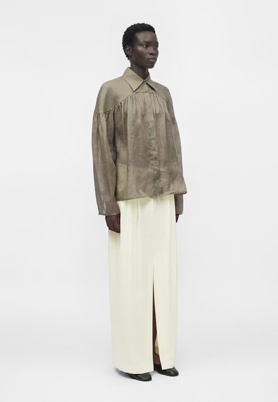 Μοντέλο με σκουρόχρωμο δέρμα φοράει φαρδιά πουκάμισα ταυπέ με μακριά μανίκια και κρεμ φούστα με ψηλή μέση και σκίσιμο μπροστά, στέκεται μπροστά σε απλό γκρι φόντο.