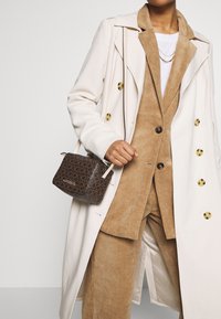 Completo in corduroy beige con t-shirt bianca, indossato sotto un trench coat beige. Borsa a tracolla marrone con motivo. Bottoni dorati visibili.