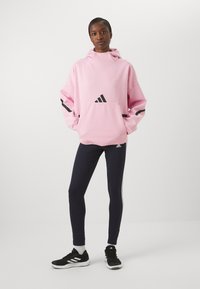 Rožinė didelis džemperis su juodais akcentais ir Adidas logotipu; derinamas su tamsiai mėlynomis Leginsais su baltomis šoninėmis juostelėmis ir juodais sportiniais bateliais.
