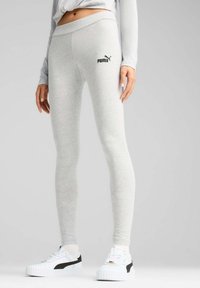 Graue Leggings aus weichem Stoff, mit einem breiten Bund und einem kleinen schwarzen Puma-Logo auf dem Oberschenkel. Kombiniert mit weißen Sneakers.