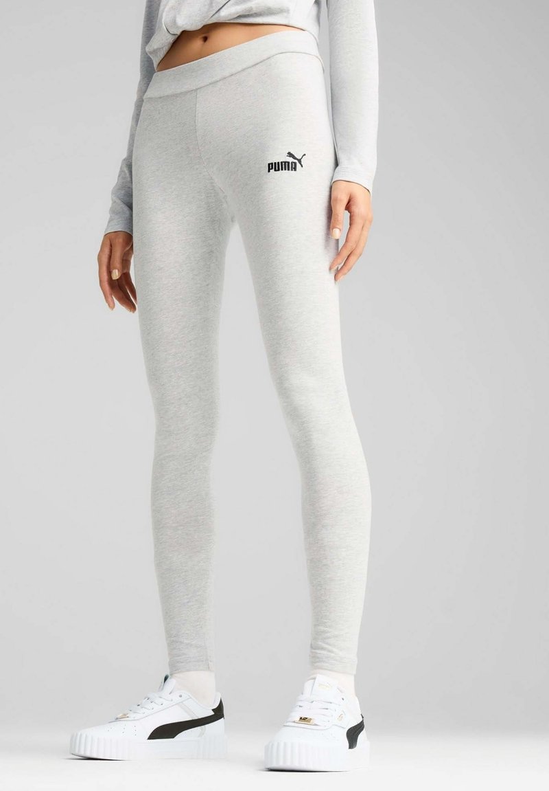 Graue Leggings aus weichem Stoff, mit einem breiten Bund und einem kleinen schwarzen Puma-Logo auf dem Oberschenkel. Kombiniert mit weißen Sneakers.