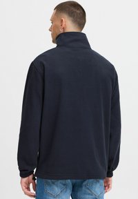 Pullover in pile di colore navy con alto colletto, maniche lunghe e polsini elasticizzati. La parte posteriore presenta una texture liscia e dettagli di cucitura minimi.