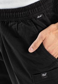Bermudas de algodón negro con cintura elástica, bolsillo frontal y etiqueta del logo de la marca visible en el lateral. Textura suave y diseño minimalista.