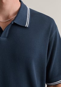 Granatowa koszulka polo z teksturowanej tkaniny, z klasycznym kołnierzykiem i cienkimi białymi paskami wzdłuż krawędzi. Krótkie rękawy.