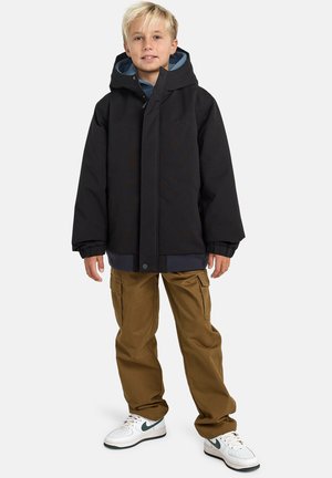 Schwarze Kapuzenjacke mit einem vorderen Druckknopfverschluss über einem blauen Hoodie, kombiniert mit beigen Cargo-Hosen und weißen Sneakern mit grünen Akzenten.