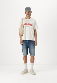 Camiseta clara con logo rojo, pantalones cortos de mezclilla azul, gorra gris, zapatillas crema, calcetines blancos y un bolso cruzado beige. Atuendo casual.