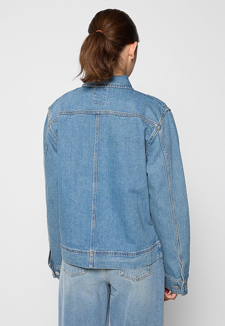 Giacca di jeans azzurro chiaro con una forma squadrata, dotata di colletto a punta, maniche lunghe e polsini con bottoni. Semplice dettaglio della cucitura posteriore.