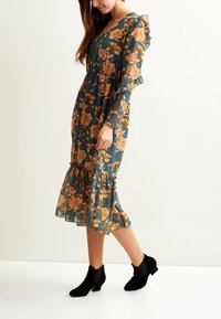 Abito midi floreale con sfondo teal, caratterizzato da fiori gialli e rosa, maniche arricciate e orlo a strati. Abbinato a stivaletti neri.