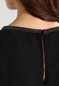Schwarze Bluse mit einem bestickten Ausschnitt, öffnung am rückwärtigen Schlüsselbereich und weicher, glatter Textur.