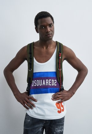 Jeune homme les mains sur les hanches portant un débardeur blanc à rayures bleues et violettes, numéro 95, et un sac à dos vert olive sur fond uni.