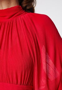 Rode plissé jurk met hoge hals, gedrapeerde mouwen en textuurstof. Beschikt over zijdezachte, vloeiende lijnen en een getailleerde taille.