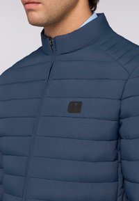 Marinfärgad quiltad jacka med hög krage, dragkedjestängning och en liten svart logotyp på bröstet med horisontell sömdekoration.