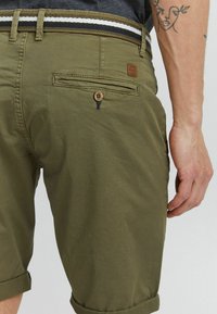 Shorts vert olive avec un ourlet roulé, dotés d'une patch en cuir et d'un bouton décoratif sur la poche arrière, complétés par une ceinture rayée.