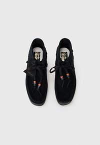 Scarpe in suede nere con punta arrotondata, cuciture a contrasto e lacci decorativi con perline colorate. La suola è durevole e testurizzata.