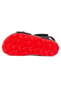 Sandale noire avec un dessus texturé et des sangles ajustables, présentant une semelle extérieure en caoutchouc rouge vif avec un motif circulaire.