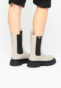 Botines de cuero beige con paneles elásticos negros, punteras redondas y suelas gruesas de goma negra. Cuentan con pestañas para facilitar su uso.