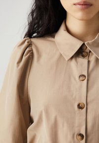 Närbild av en kvinnas överkropp klädd i en ljusbrun skjorta med knappar och puffärmar, med en klassisk krage och knappar.
