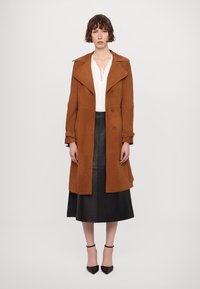 Trench coat in suede marrone con colletto oversize, maniche strette e bottoni doppi; abbinato a una gonna in pelle nera e tacchi.