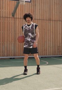 Jugador de baloncesto vistiendo una camiseta sin mangas de patrón gris y blanco, pantalones cortos negros y zapatillas negras, sosteniendo un balón de baloncesto en una cancha.