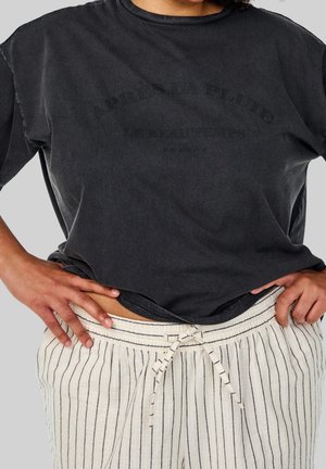 Persoon draag een loszittend zwart T-shirt en witte broek met koordje en zwarte verticale strepen, handen rustend op de heupen.