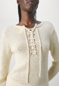 Pull en tricot crème avec un devant à lacets et oeillets argentés, texture douce et encolure large, présentant un design décontracté.