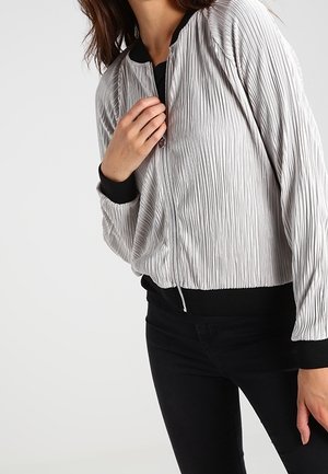 Vrouw draagt een lichtgrijs, geplooid bomberjack met zwarte manchetten en kraag, gecombineerd met zwarte broek, tegen een effen achtergrond.