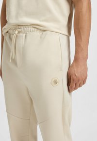 Helle beige Sporthosen mit elastischem Bund, Kordelzug und einem sechseckigen Logo am Oberschenkel. Glatter, strukturierter Stoff mit Nahtdetails.