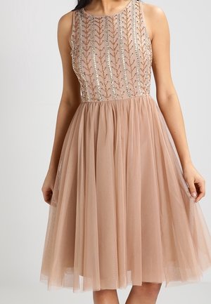 Beżowa sukienka midi bez rękawów z przejrzystą tiulową spódnicą i cekinowym gorsetem w kwiatowy wzór na stojącej kobiecie.