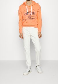 Felpa arancione con stampa frontale, pantaloni bianchi e sneakers bianche con dettagli neri. Design casual con vestibilità comoda e cappuccio con lacci.