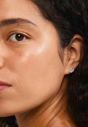 Silver stud earrings with a circular design, featuring inset clear crystals, worn by a person with curly hair and smooth skin, close-up view.