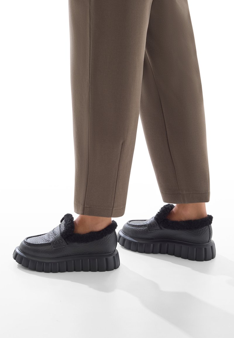 Persona che indossa scarpe slip-on nere con suole spesse e foderate in pelliccia, abbinate a pantaloni marroni a 3/4 su uno sfondo bianco.