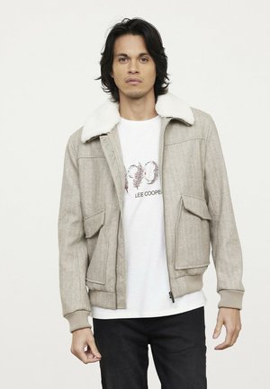 Beige wollen blend jas met een witte neplederen kraag, twee voorzakken en ribgebreide manchetten, gedragen over een witte graphic t-shirt.