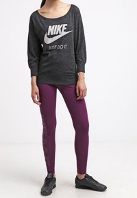 Graues Langarm-Nike-Oberteil mit weitem Ausschnitt und weißem Logo, kombiniert mit lila Leggings mit "JUST DO IT"-Druck, getragen mit schwarzen Sneakers.
