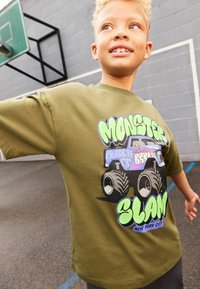 Olíva zöld pamut póló, színes monster truck grafikával, nagy kerekekkel és merész "MONSTER SLAM" felirattal kék és zöld színben.