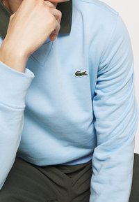 Ljusblå sweatshirt med grön krage, prydd med en broderad krokodil-logotyp på bröstet. Slät textur och avslappnad passform.