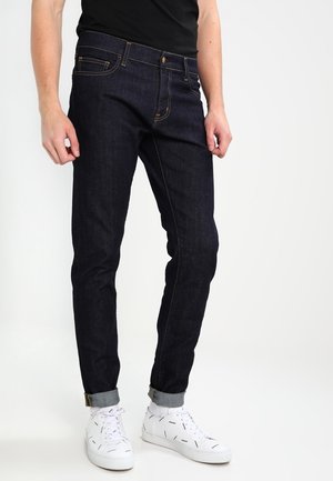 Mann trägt dunkelblaue hochgekrempelte Jeans, weiße Sneakers mit schwarzen Zickzackmustern und ein schwarzes Hemd, steht vor weißem Hintergrund.