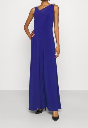 Długa wieczorowa sukienka z żywego niebieskiego materiału, z dekoltem w serek, asymetrycznym krojem na ramieniu i drapowanymi plisami; zestawiona z czarnymi szpilkami.