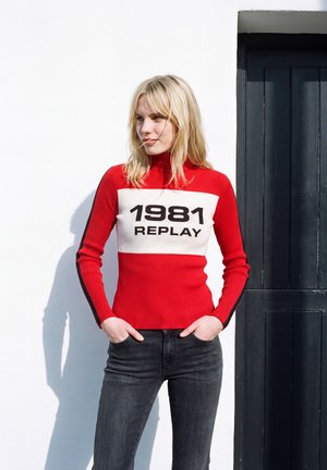 Un mannequin blond portant un pull rouge avec l’inscription « 1981 Replay » et un jean foncé se tient près d’une porte sombre, les mains dans les poches.