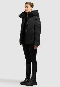 Schwarze Daunenjacke mit Chevron-Muster, hohem Kragen, Reißverschluss und Seitentaschen, kombiniert mit schwarzen Leggings und kniehohen Stiefeln.