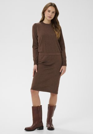 Woman standing wearing a brown long-sleeve knit dress and brown mid-calf leather boots against a plain light background.