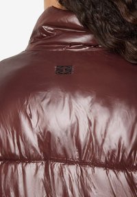 Ruda pūkinė striukė su blizgia tekstūra, aukšta apykakle ir mažu juodu logotipu nugaroje. Matomas siuvimo siūlės per siūles.